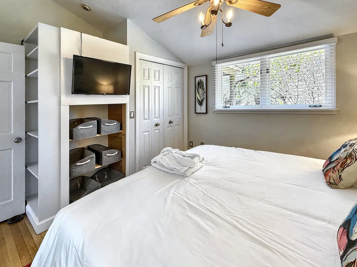 A large, neatly made bed faces a wall-mounted TV, complemented by a window allowing natural light to fill the room. Decorative plant-themed artwork provides a soothing backdrop, while the overall setup is designed for lounging and entertainment.