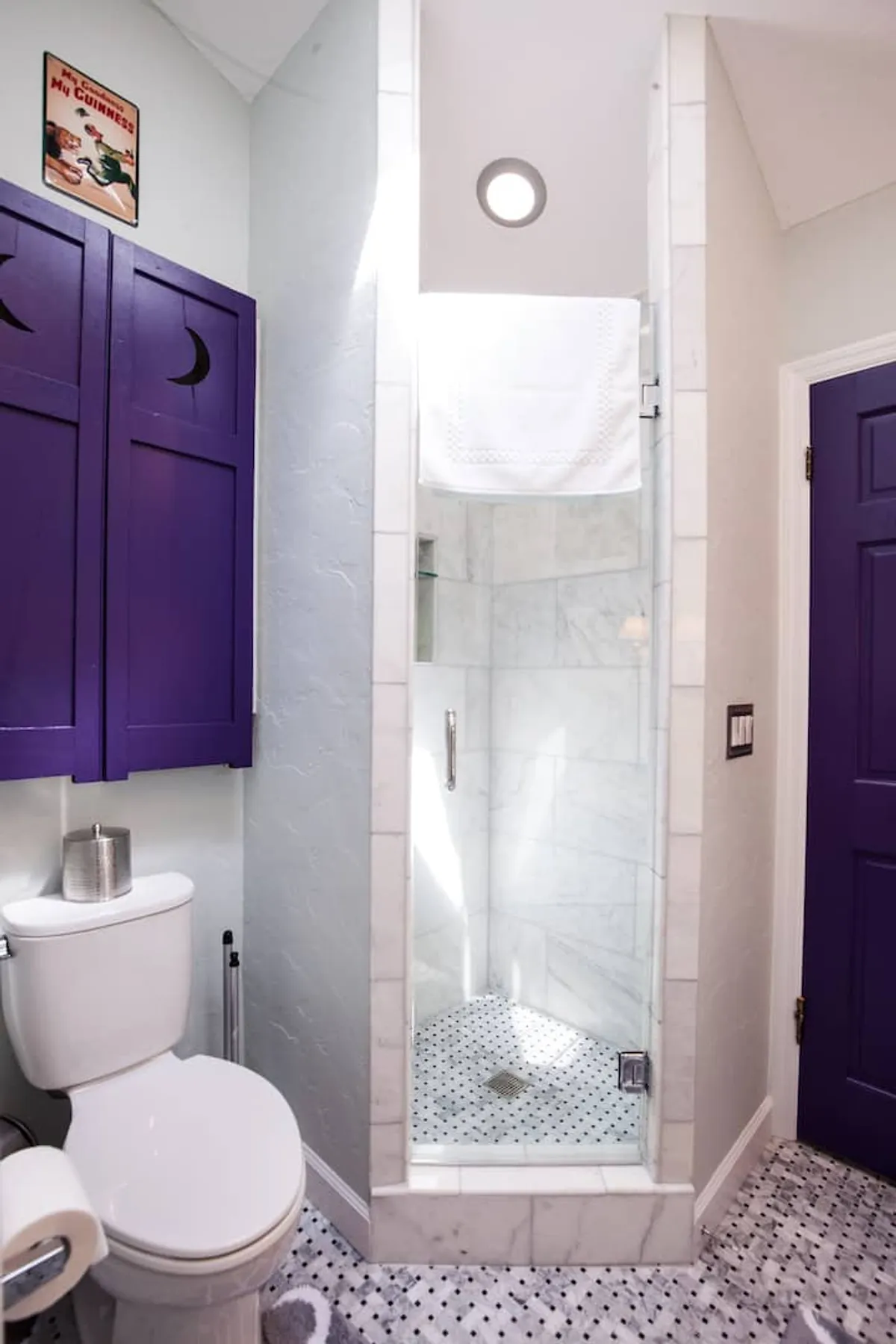 A contemporary bathroom with purple cabinet doors, a glass shower enclosure, marble shower tiles, and decorative wall art. The room exudes a playful, modern vibe while offering a spacious, refreshing shower experience.