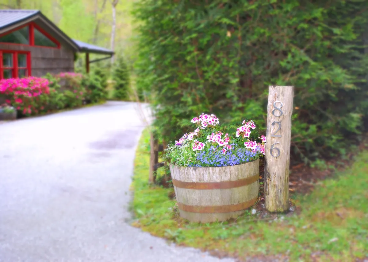 The image features the home's inviting exterior, with a charming flower planter, a welcoming wooden sign by the entrance, and lush foliage suggesting proximity to gardens and walkable outdoor spaces. The scene is alive with the possibility of enjoying the garden, taking a leisurely walk, or greeting guests, all set in a tranquil, residential neighborhood.