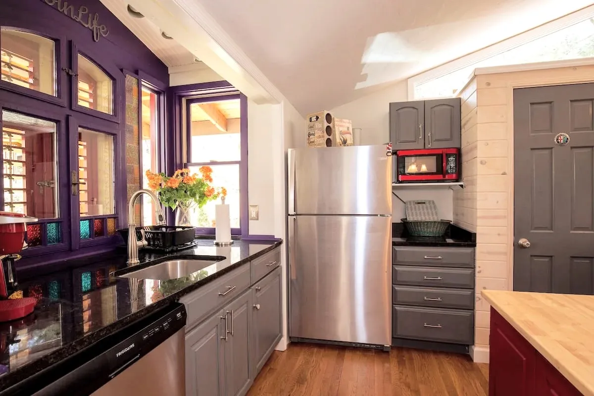 A kitchen space with distinctive purple cabinetry, stainless steel appliances, and sunlight streaming through large windows. A floral arrangement adds a festive touch, and the space feels lively and personal—ideal for hosting friends or enjoying a morning cup of coffee.