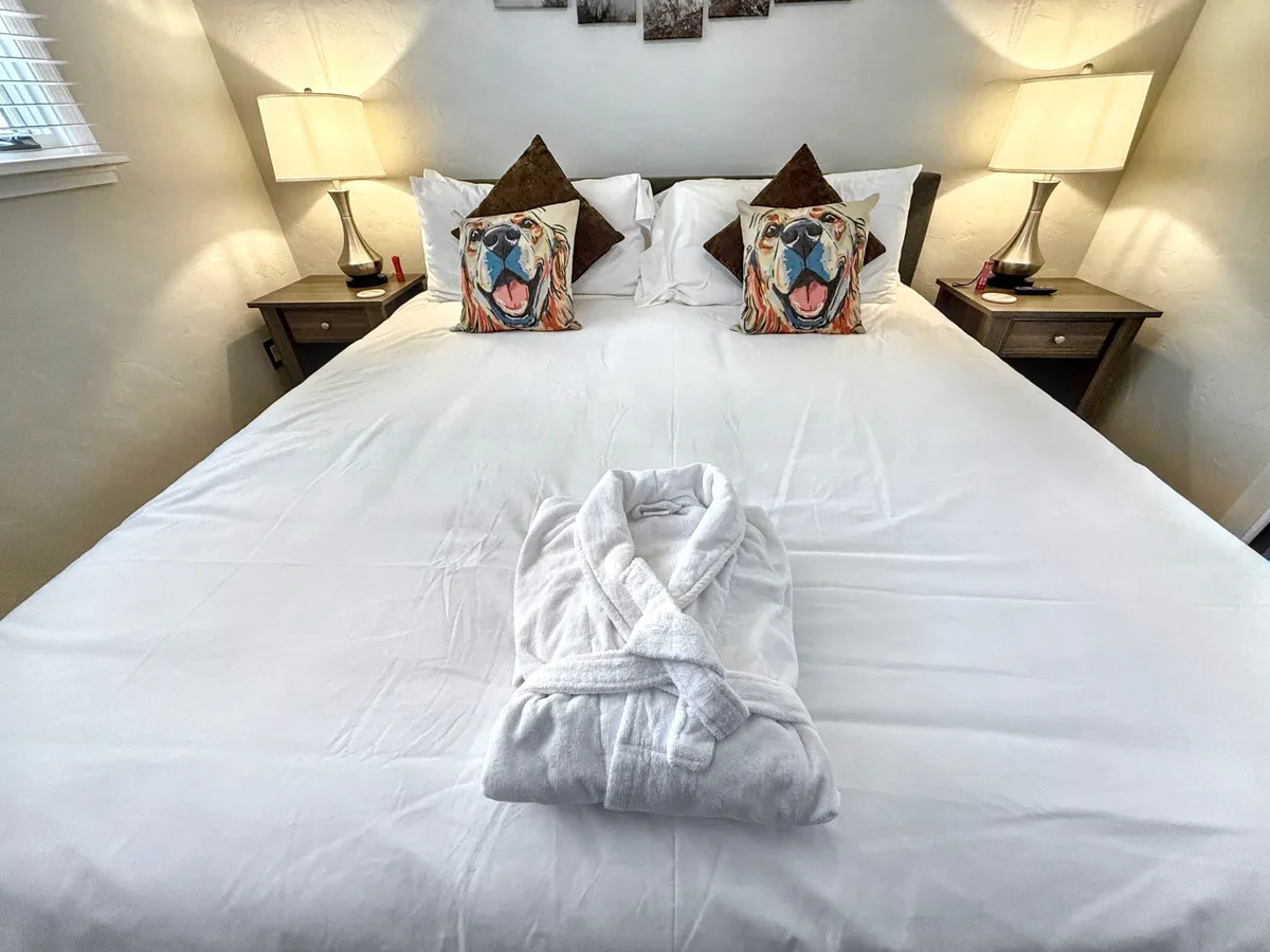 A plush bed topped with white linens, playful decorative animal-themed pillows, and a soft white robe draped across the bed. Lamps on either side of the bed and a cozy atmosphere suggest comfort and luxury.