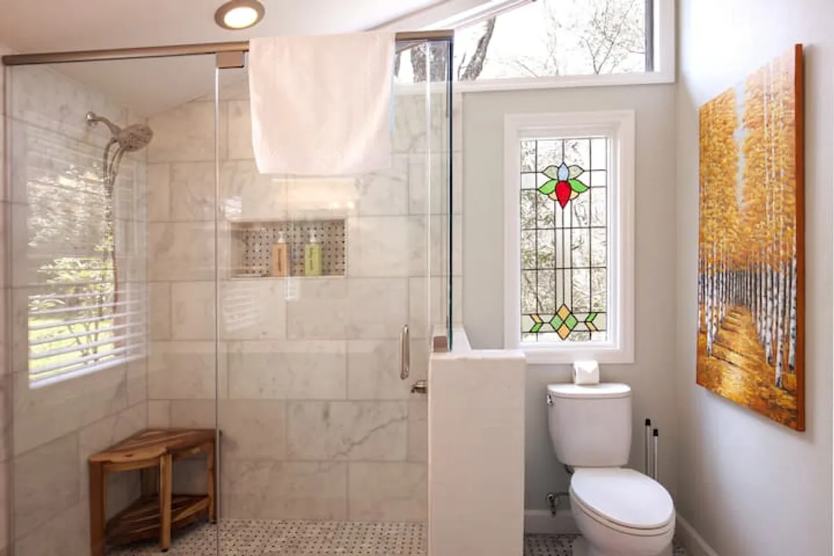 A bright, serene bathroom featuring a glass shower enclosure, a stained glass window casting colorful light, a wooden stool, and vibrant artwork. The space is filled with natural light, creating a peaceful atmosphere ideal for relaxation after a day outdoors.
