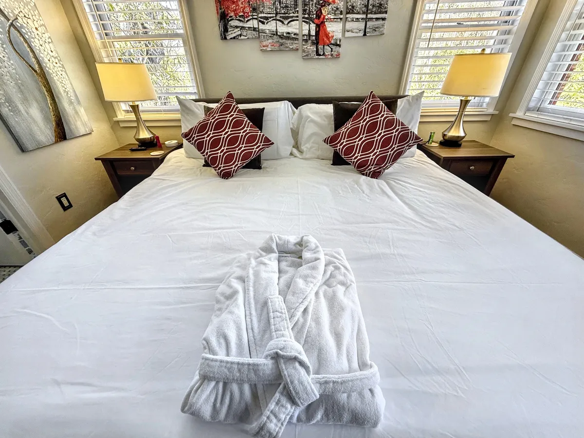The bed is topped with crisp white linens and a plush bathrobe, with decorative pillows and tasteful artwork adding a boutique touch. A lamp on the nightstand creates a soft, inviting glow—suggesting a peaceful setting for relaxation.
