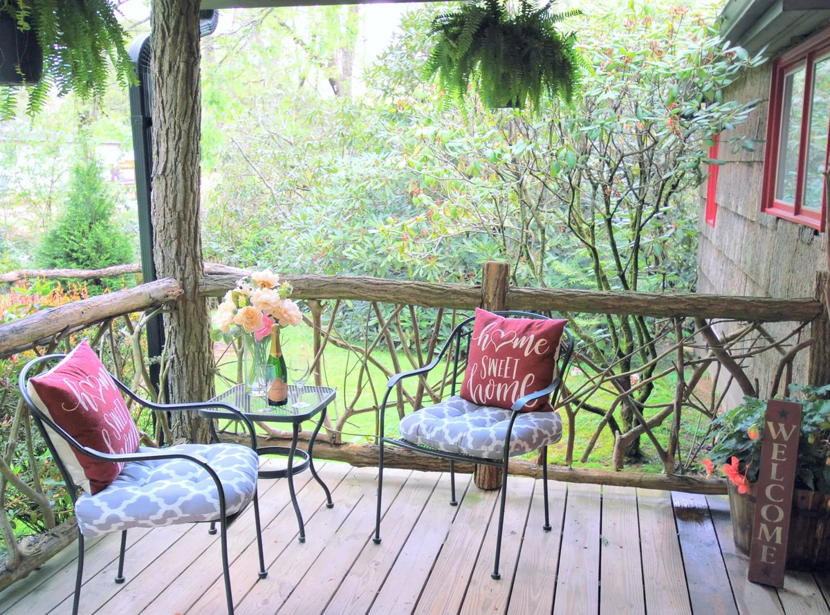 A cozy porch with a rustic wooden railing, a small table adorned with flowers in a vase, and decorative pillows invites relaxation. The setting suggests early morning tranquility or festive gatherings, offering a transitional space between the home and the vibrant town just beyond.