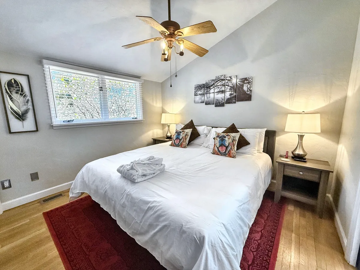 A large king bed with crisp white linens and decorative pillows anchors the room. Nightstands with lamps flank either side, with wall art adding a homey touch. Sunlight gently brightens the space, emphasizing the inviting and serene atmosphere.