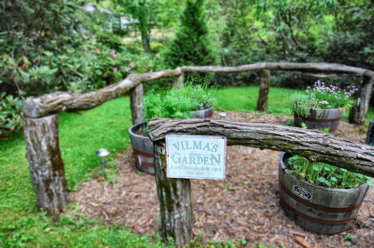 A charming garden area bordered by a wooden fence, featuring herb planters and a welcoming garden sign. The peaceful setting is ideal for gardening, leisurely strolls, or simply enjoying the scents and colors of nature.