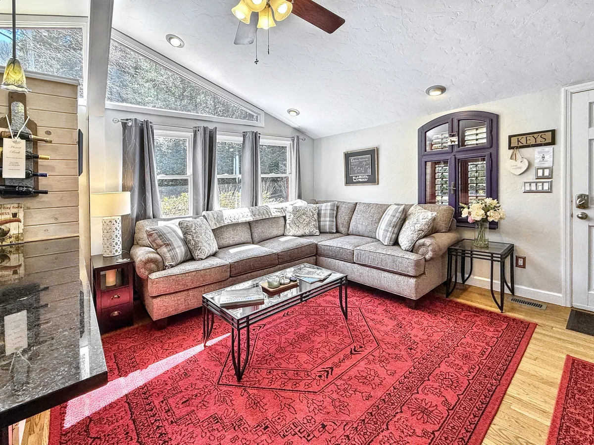 The image features a large, ultra-comfortable sectional sofa, a vibrant red area rug, a coffee table, and expansive windows letting in natural light. The scene exudes coziness and is ideal for gathering for movie nights, board games, or simply sharing stories.