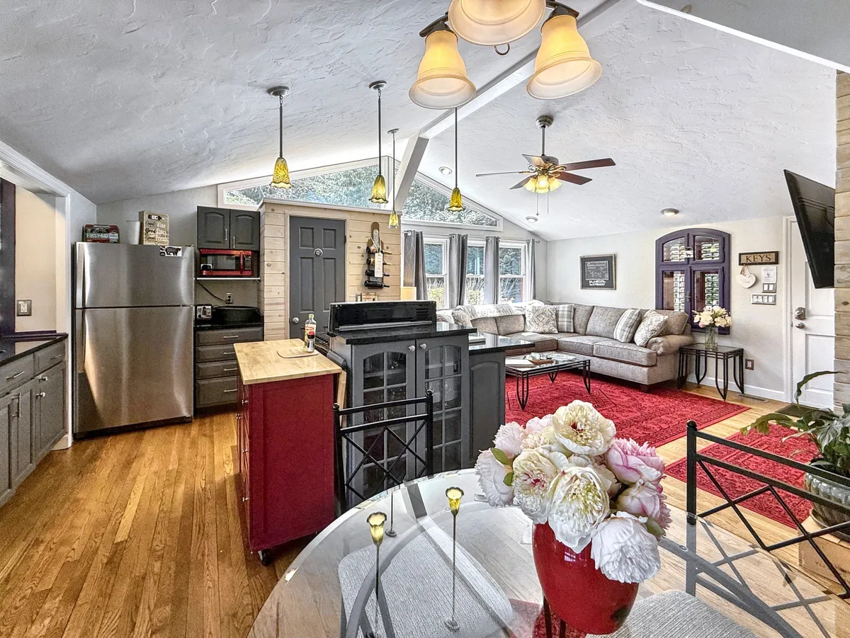 This image showcases an inviting open-concept space featuring a kitchen island, a comfortable sofa, a dining table adorned with flowers, and large windows that bathe the room in natural light. The arrangement emphasizes social interaction and flexibility, reflecting a space where cooking, dining, and relaxing happen seamlessly together.